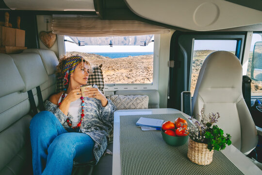 Cute trendy adult woman have relax leisure activity laying on the sofa inside her cozy alternative home camper van. Concept of tourism and modern vanlife. Beach and ocean view outside the window
