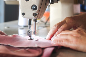 Hand of seamstress using industrial sewing machine to sew the seams of pink cloth close-up.