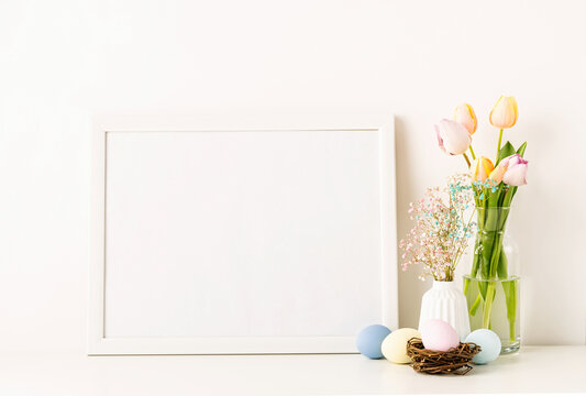 Mockup with a white frame spring tulips in vase, gypsophila and pastel colored eggs on light background