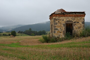 Petite maison de vigneron en pierre dans un  champ avec à l’arrière un banc de brouillard descendant de la montagne 