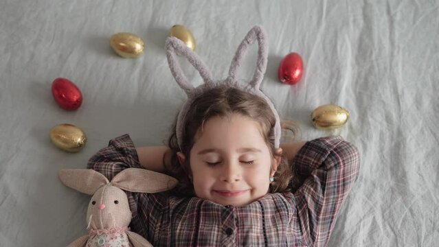 Adorable little girl with gray bunny ears sleeping in bed at home with chocolate Easter eggs and bunny toy. Easter greeting card. Easter Bunny kid