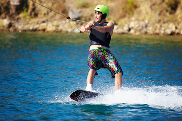 A young man wearing a helmut and lifejacket wakeboarding on a lake. A young man wearing a helmet and lifejacket wakeboarding on a lake. - obrazy, fototapety, plakaty