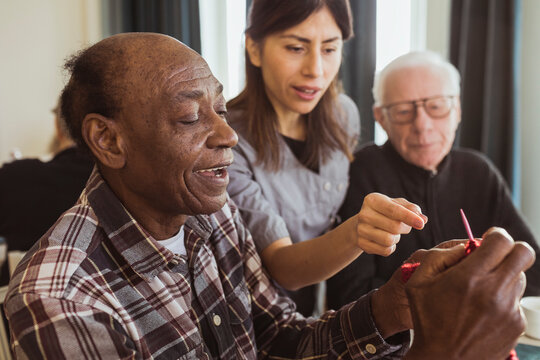 Female caregiver teaching senior man knitting in nursing home