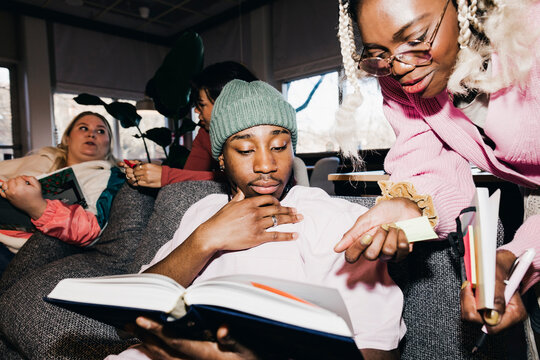 Male and female discussing over book while friends in background at college dorm
