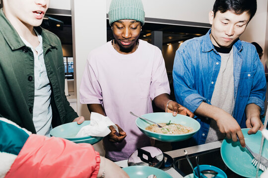 Young man with noodles while roommate cleaning dishes at sink in college dorm