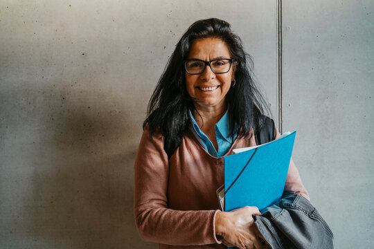 Smiling mature woman with books and file standing against gray wall at university
