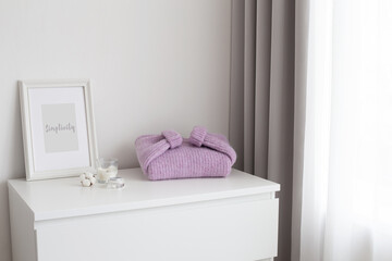 Frame with text SIMPLICITY, lilac knitted sweater and candles stand on white chest of drawers. Spring bedroom home decor. White stylish interior.