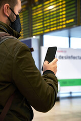 Young man with mask at the airport looking at flight schedule through mobile