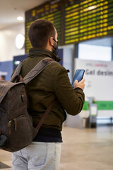 Young man with mask at the airport looking at flight schedule through mobile