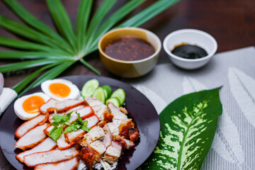 Steamed rice and red pork, crispy pork in a black plate Served with dipping sauce and dark soy sauce Place it on a black wooden table.