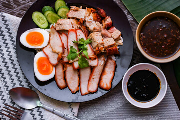 Steamed rice and red pork, crispy pork in a black plate Served with dipping sauce and dark soy sauce Place it on a black wooden table.