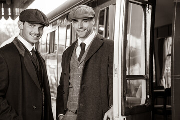 Handsome English gangsters standing next to train at railway station, laughing, with train in background - obrazy, fototapety, plakaty