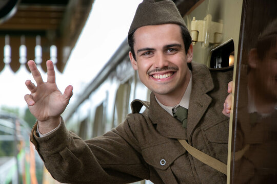 Handsome male British soldier in WW2 vintage uniform at train station leaning out of train, waving and smiling