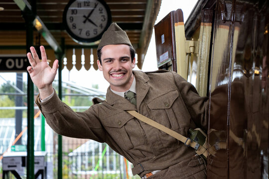Handsome male British soldier in WW2 vintage uniform at train station leaning out of train, waving and smiling