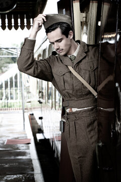 Handsome pensive male British soldier in WW2 vintage uniform at train station standing on train, putting on hat