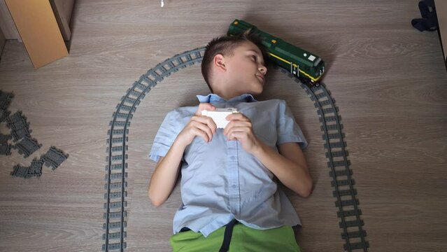 a teenager is playing with a train on the controls on the floor in the room. top view. toy railway. smiling, he turns his head to the moving locomotive. there is light from the window in the room