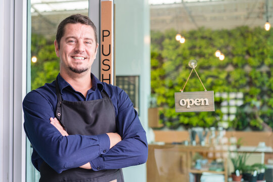 Portrait of male barista owner in apron smiling and standing on front of cafe with open sign board in coffee shop