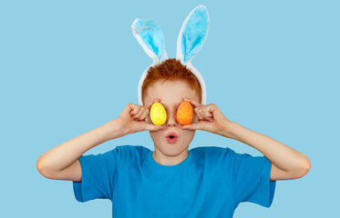 Portrait of a surprised red-haired boy in rabbit ears, who holds colorful Easter eggs in front of...