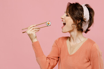 Side view young caucasian fun woman 20s wearing casual clothes hold in hand eat biting with chopsticks makizushi sushi philadelphia roll japanese food isolated on plain pastel pink color background