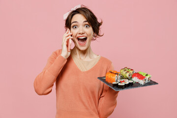 Young surprised amazed shocked woman in sweater hold in hand makizushi sushi roll served on black plate traditional japanese food talk on mobile cell phone isolated on plain pastel pink background