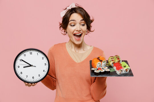 Young excited amazed happy fun cheerful woman 20s wear casual clothes hold in hand makizushi sushi roll served on black plate traditional japanese food clock isolated on plain pastel pink background