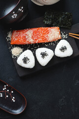 Japanese onigiri and slice of salmon fillet on a black wooden chopping board, high angle view on a black stone surface, vertical shot with space
