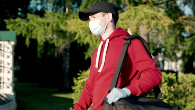 Asian delivery man in red uniform with face mask carrying bag of food delivering food to customer home during covid virus pandemic. He walking outdoor in village looking for customer home address