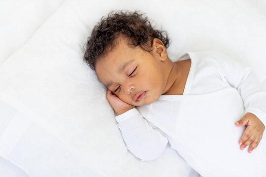 African-American little baby sleeps on a white bed at home with her hand folded under her cheek