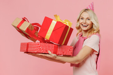 Side view elderly happy smiling satisfied woman 50s in t-shirt birthday hat hold red present box with gift ribbon bow isolated on plain pastel pink background studio Celebrating party holiday concept.
