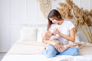 a mother with a baby in her arms feeds milk from a bottle on the bed at home in a bright room, the...