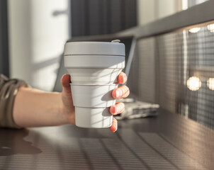 Woman hand closeup holding takeaway eco collapsible cup. Student or freelancer sitting at table...