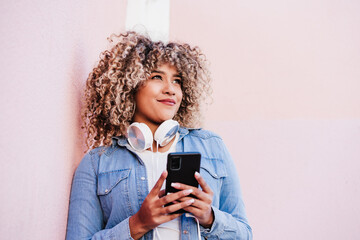 portrait of smiling hispanic woman with afro hair in city using mobile phone and headset. lifestyle