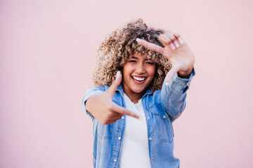 portrait of beautiful hispanic woman afro hair outdoors in spring doing frame with hands. pink wall