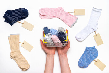 Female hands holding shopping basket with socks among women's cotton socks set with price tags on white background. Fashionable socks store. Socks shopping, sale, merchandise, advertisement concept