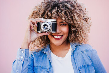 beautiful happy hispanic woman with afro hair holding vintage camera. pink background