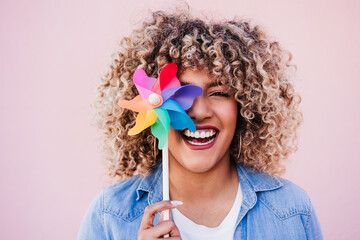 beautiful happy hispanic woman with afro hair holding colorful pinwheel. pink background,wind energy - 489560394