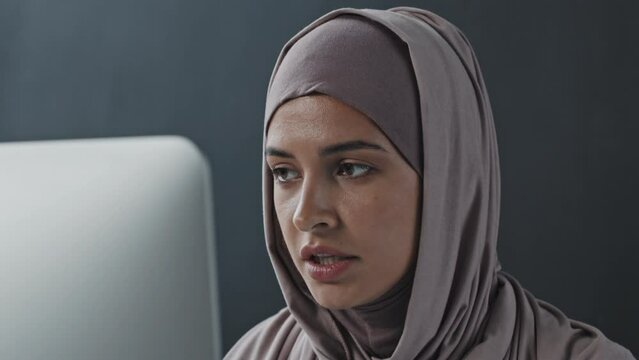 Chest up of young Muslim woman in hijab talking on online video call on computer in office