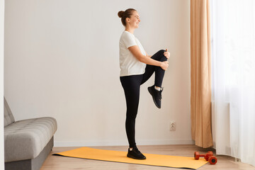 Obraz premium Side view full length of sporty woman wearing white t shirt and black leggins doing sport exercises at home, standing with raised leg, looking ahead, posing near sofa in front of window.