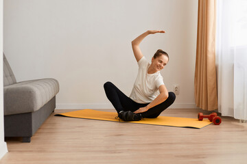 Woman wearing white t shirt and black leggins doing sport exercises at home, sitting in lotus position on mat practicing yoga, bending to side with raised hand, doing stretching workouts.