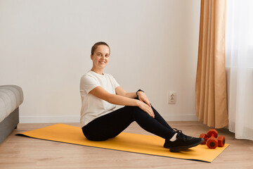 Full length shot of attractive sporty woman wearing white t shirt and black leggins doing sport exercises at home, sitting on floor and looking at camera with happy positive expression. © sementsova321