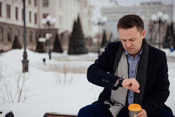 A young man in a coat and suit sits on a bench in a city park looking at his watch with a cup of coffee