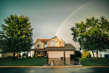 A young girl plays on her driveway in front of her house with a rainbow behind it.