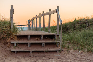 Fototapeta premium An old wooden path and stairs built over beach grass with water in the background during sunset.