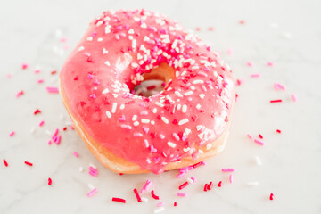 A donut with pink frosting and Valentines pink, white, and red heart sprinkles on a white marble background