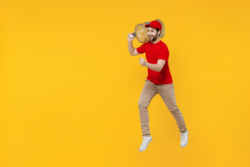 Professional side view fun delivery guy employee man in red cap T-shirt uniform workwear work as dealer courier hold water bottle jump high isolated on plain yellow background studio. Service concept.