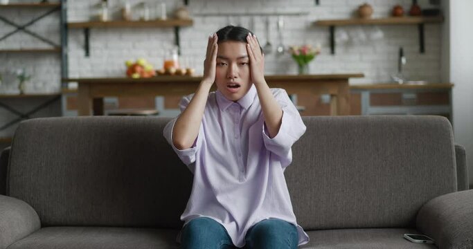 Scared asian woman shocked. Attractive young female unpleasantly surprised looking at the camera while sitting on sofa at home. She covers her mouth with her hand of modern kitchen background