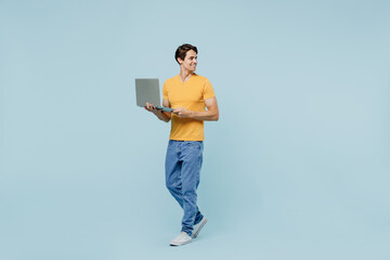 Full body young man 20s wear yellow t-shirt hold use work on laptop pc computer look aside on workspace area mock up isolated on plain pastel light blue background studio. People lifestyle concept