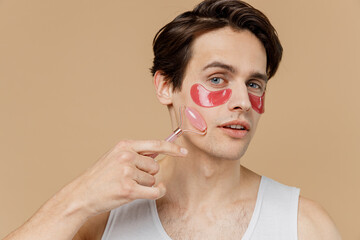 Young man 20s perfect skin wear pink patch under eye do face massage with jade facial roller quartz...