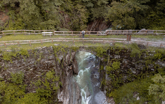 Aerial photo of a photographer taking a picture from an old stone bridge, surrounded by lush vegetation. An emerald-colored river passes beneath the bridge, on a rocky riverbed carved out by water and