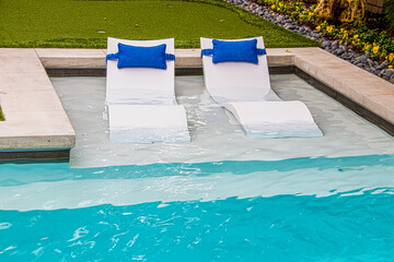 Comfy white loungers in shallow end of refreshing blue swimming pool in yard with flower trim around the edge